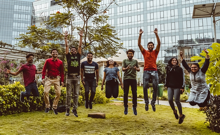 Nine people jumping mid-air outside with a building in the background.