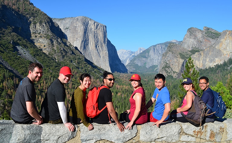 Group of people sit on rock ledge with foliage and mountains in the background.