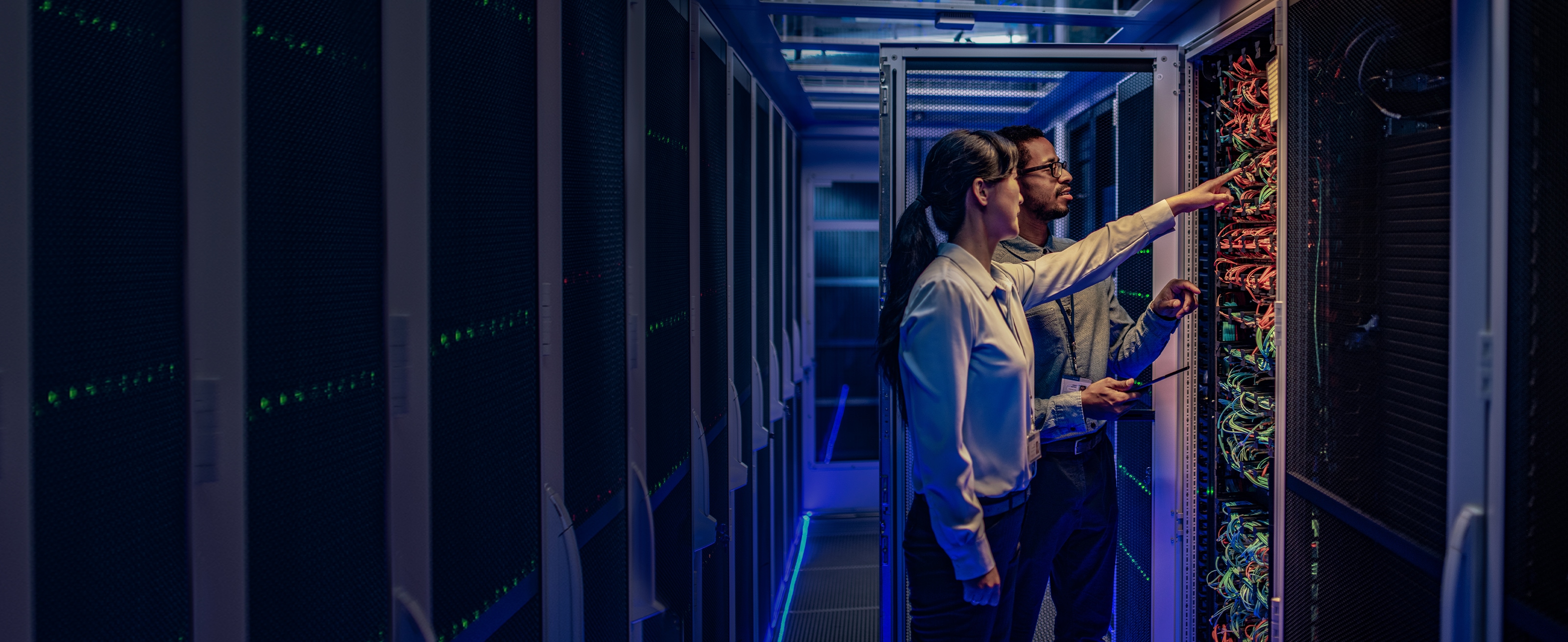 Two employees in a data center pointing at ethernet cables on the back of a rack of networking equipment. 