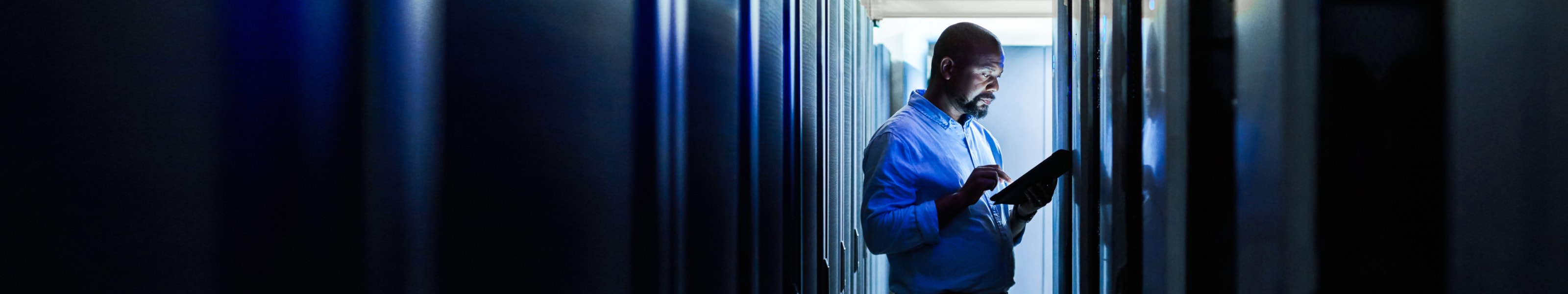 IT worker walking through the enterprise data centre checking network dashboards on a tablet.