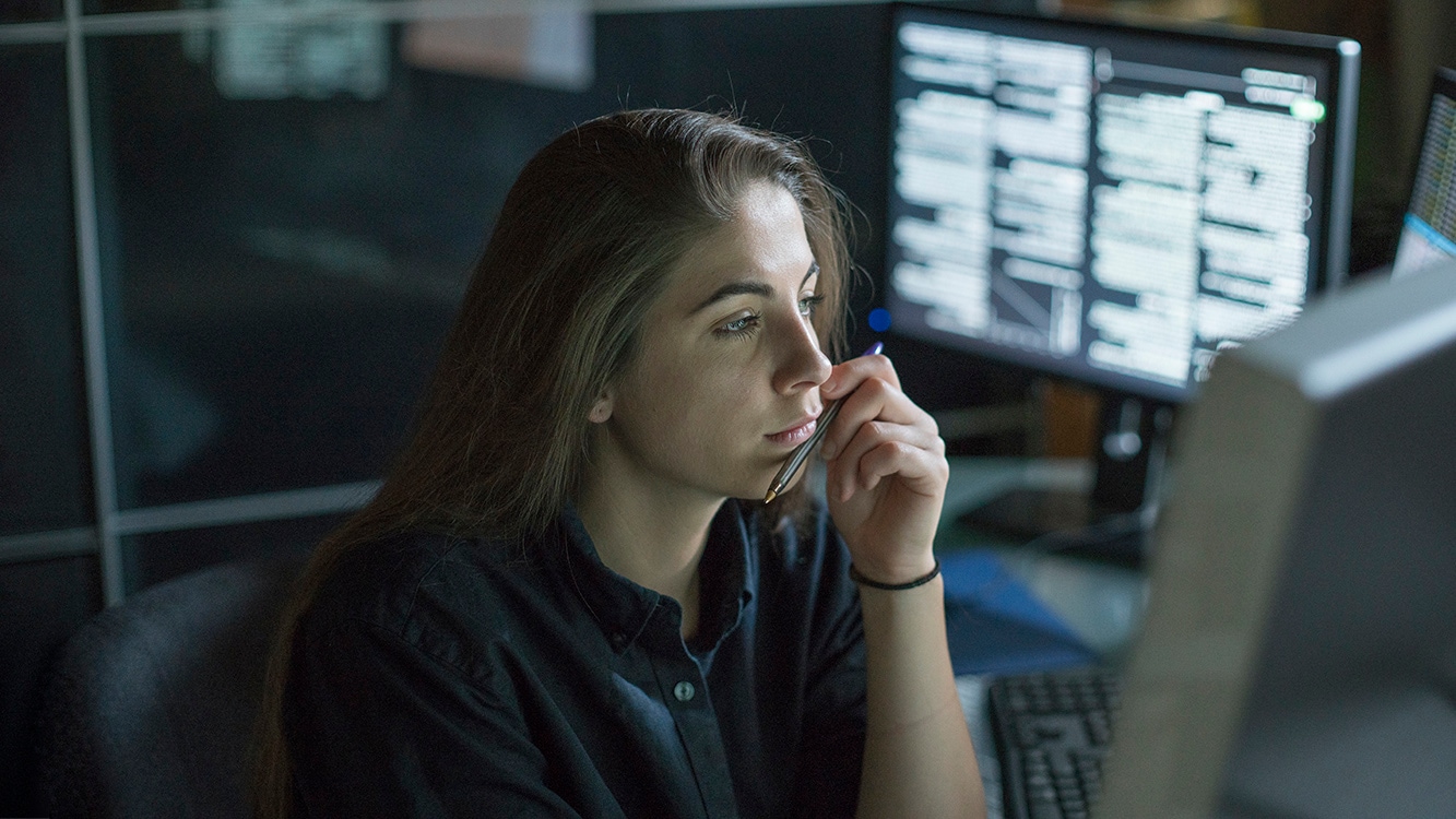 A young woman is seated at a desk surrounded by monitors displaying data, she is contemplating in this dark, moody office.